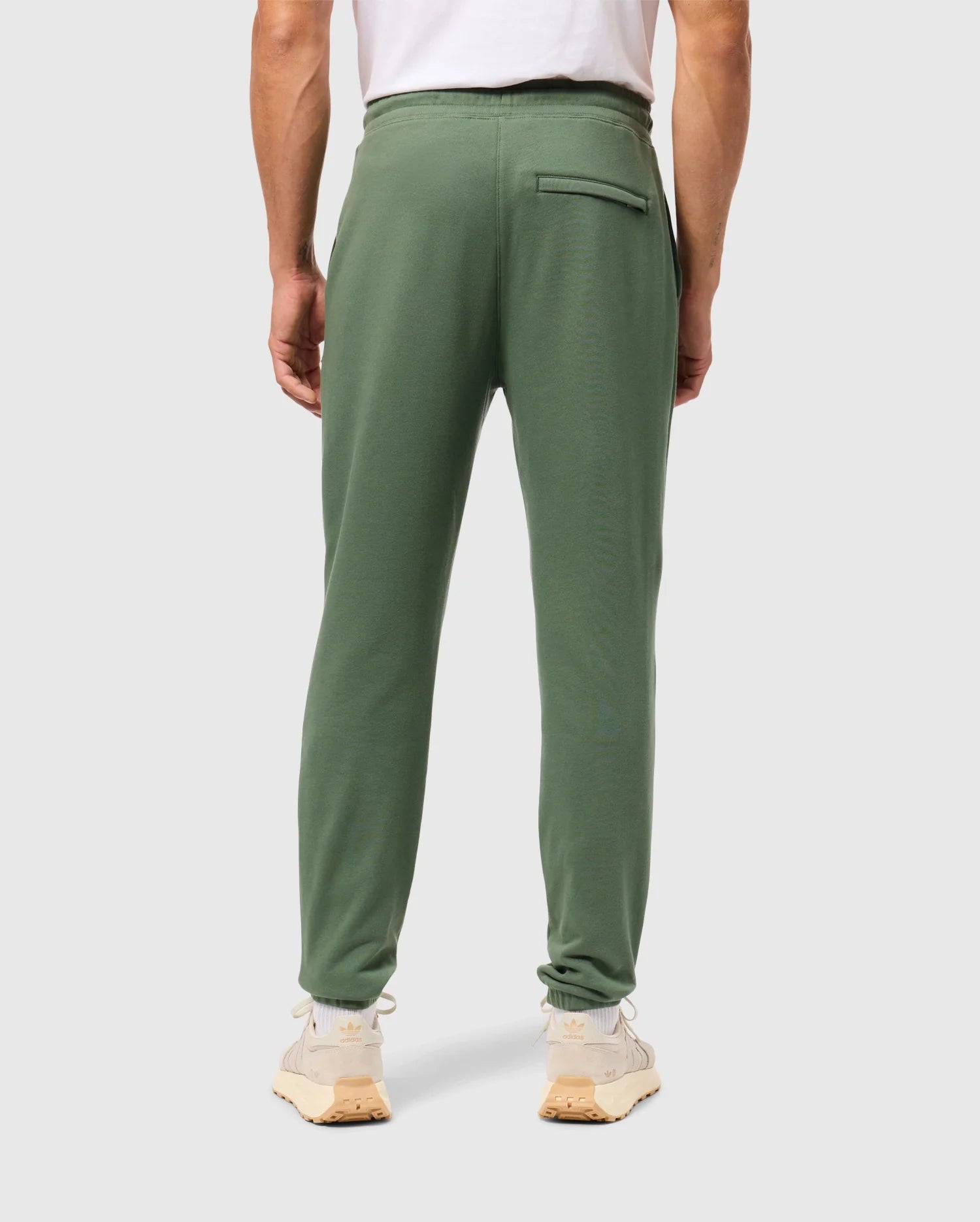 A person is wearing casual bottom Psyho Bunny Men's Pacific Chenille Joggers, paired with beige sneakers and a white t-shirt. They are standing with their back to the camera against a plain gray background.