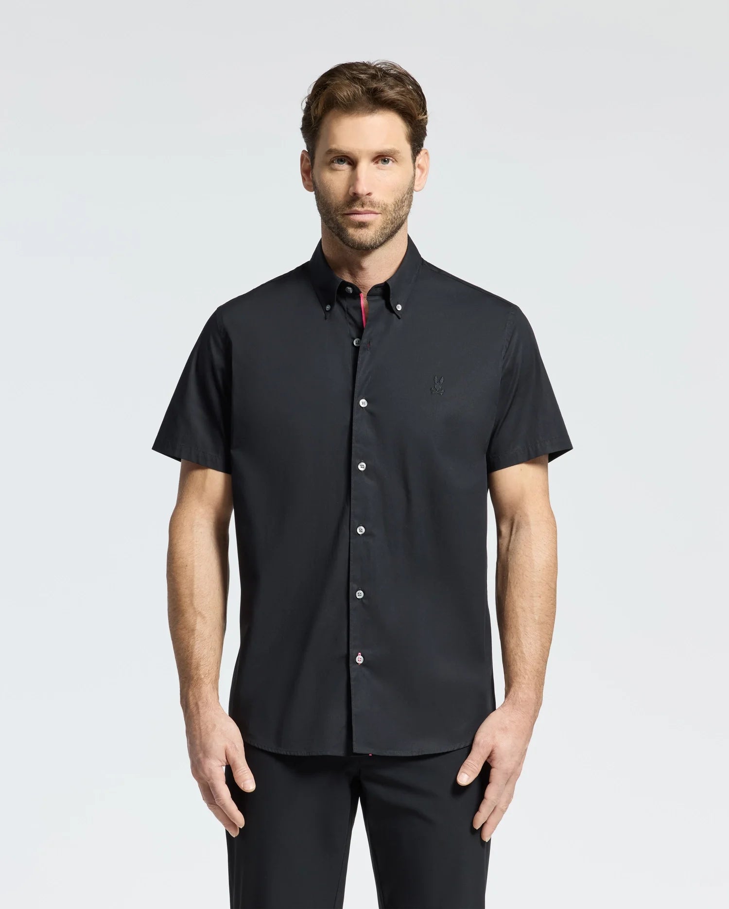 A man stands against a plain background wearing a Psycho Bunny MENS GABRIEL SHORT SLEEVE SHIRT in black, paired with matching pants. Crafted from premium Pima cotton, the ensemble complements his direct gaze and neutral expression.