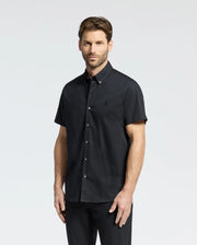 A brown-haired, bearded man in a Psycho Bunny black short-sleeve Gabriel shirt (B6Q351F200) made of woven stretch fabric stands against a white background, looking neutrally at the camera.