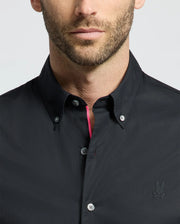 Close-up of a man wearing the Psycho Bunny MENS GABRIEL SHORT SLEEVE SHIRT - B6Q351F200, a black Pima cotton button-up. It includes an embroidered chest logo and stretch fabric for comfort, with bright pink on the inner placket against a plain, light background.