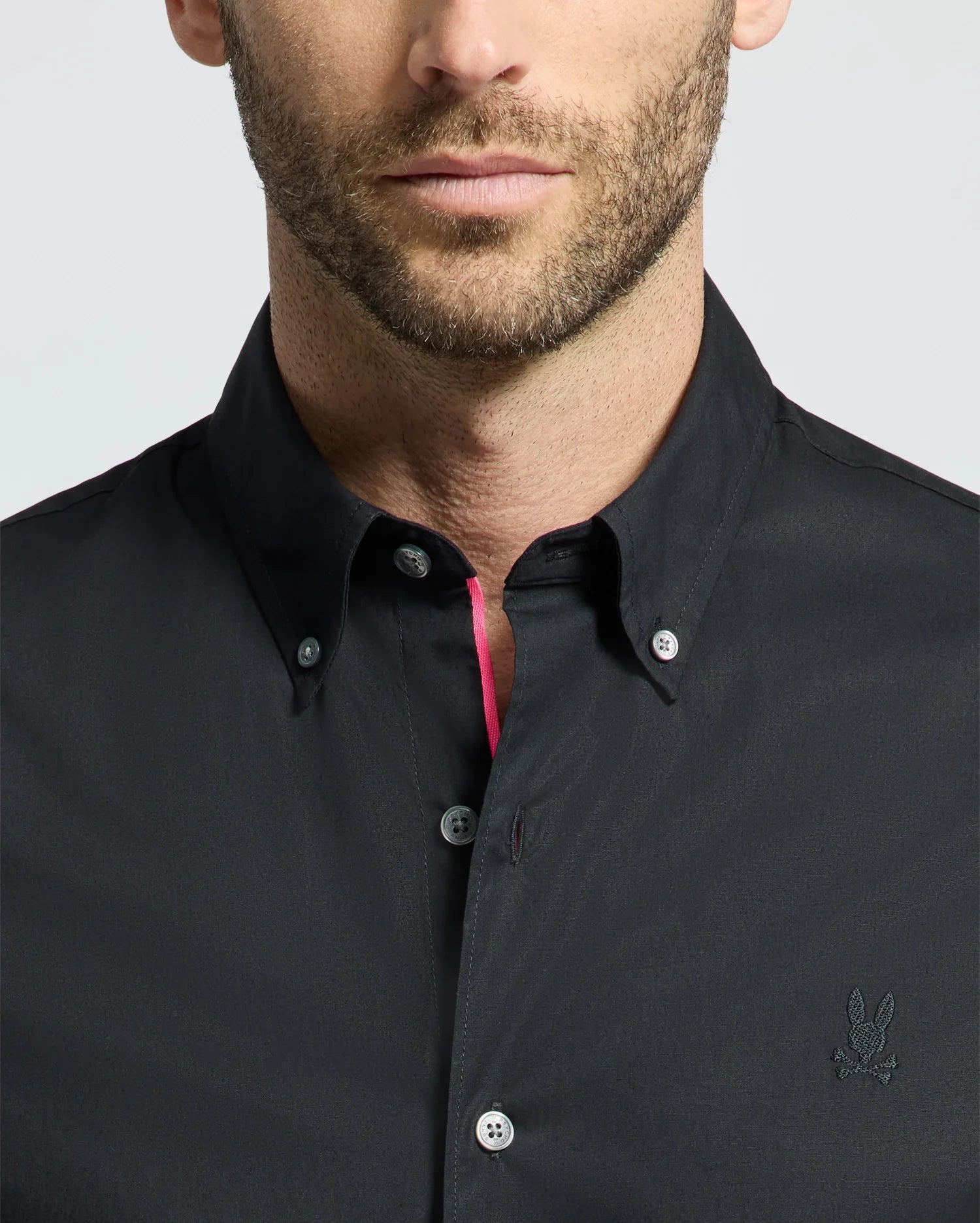 Close-up of a man wearing the Psycho Bunny MENS GABRIEL SHORT SLEEVE SHIRT - B6Q351F200, a black Pima cotton button-up. It includes an embroidered chest logo and stretch fabric for comfort, with bright pink on the inner placket against a plain, light background.