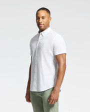 Wearing a Psycho Bunny MENS GABRIEL SHORT SLEEVE SHIRT - B6Q351F200 in white and light olive green pants, a person stands against a plain white background, looking at the camera with a slight smile.
