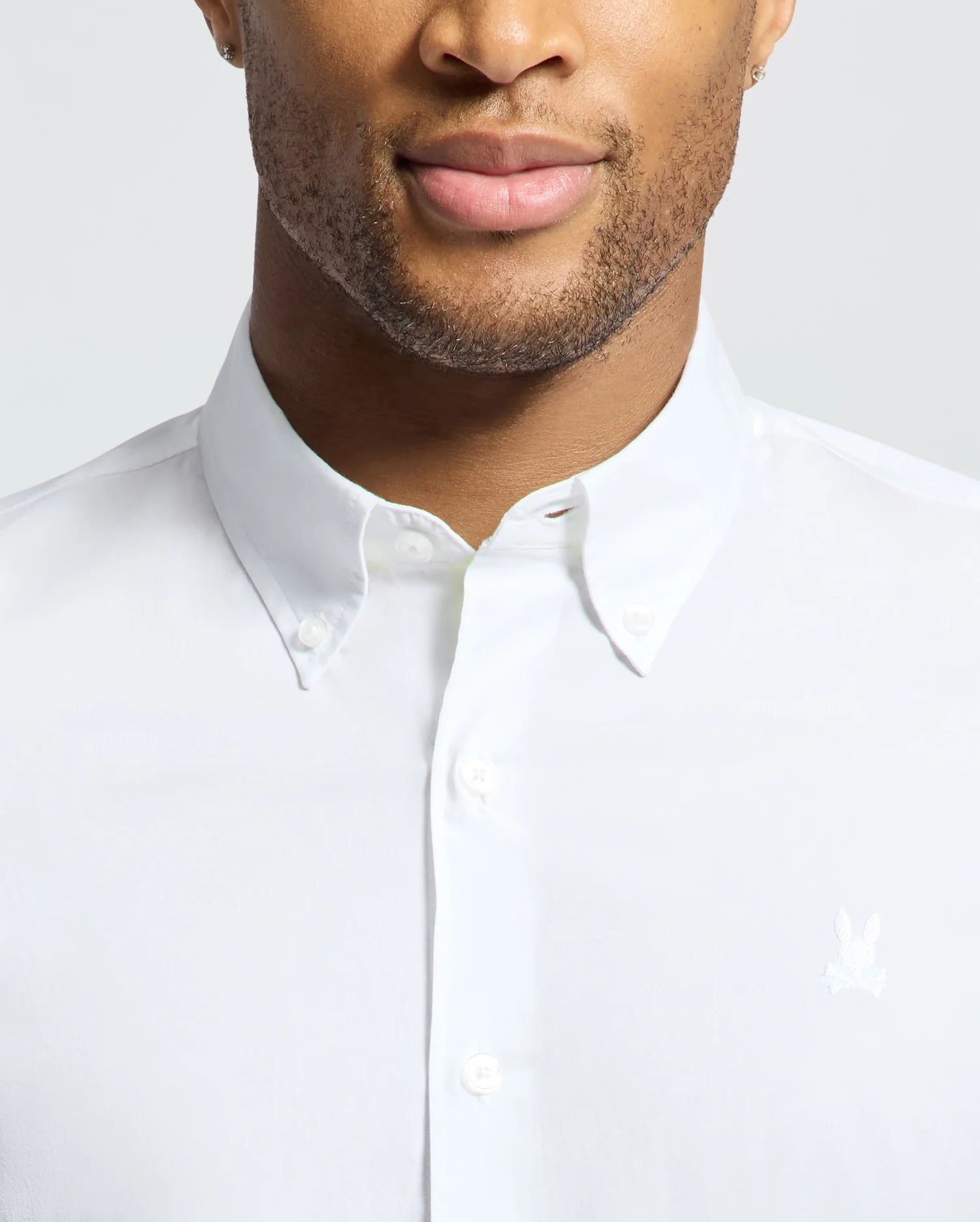 A person in a Psycho Bunny white Gabriel short sleeve shirt, made from luxurious Pima cotton with an embroidered Bunny logo on the chest. The image shows the upper torso and face with a neutral expression, featuring a neatly buttoned classic collar for a polished look.