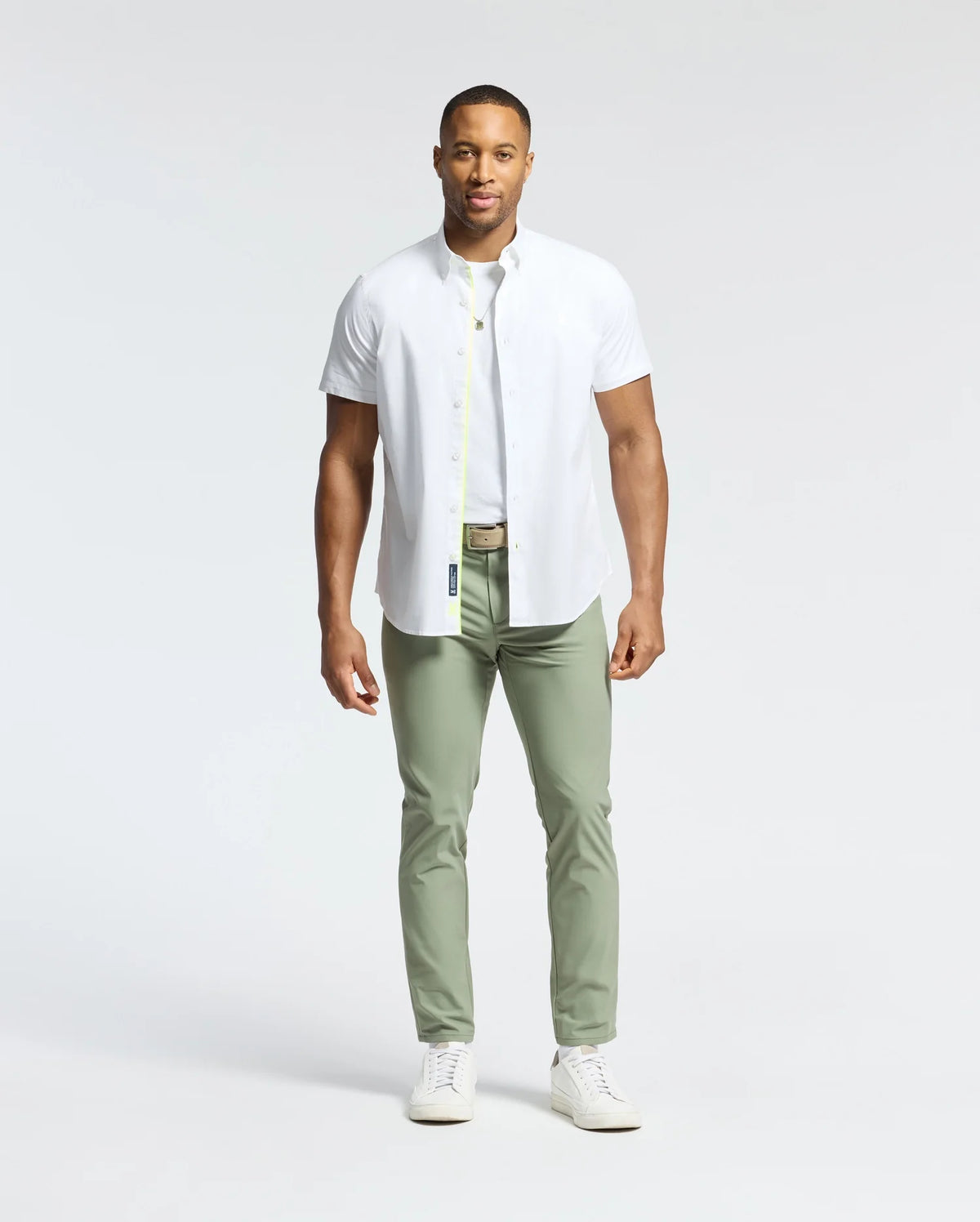 A man stands against a plain background wearing a crisp white Psycho Bunny Gabriel short sleeve shirt, complemented by light olive green pants and pristine white sneakers.