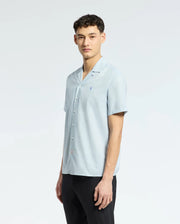 A person with short dark hair is wearing a Psycho Bunny MENS COBAR WOVEN SHIRT - B6Q522F200 and black pants, standing against a plain white background, looking toward the camera.