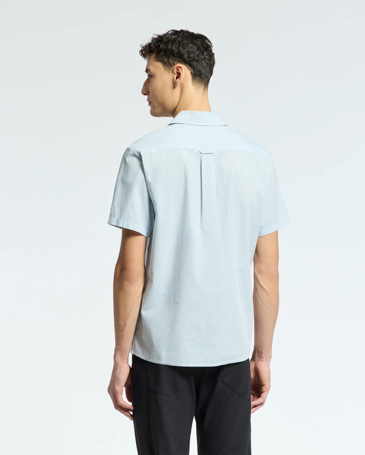 A person with short dark hair is shown from behind, wearing a light blue Psycho Bunny MENS COBAR WOVEN SHIRT - B6Q522F200 and black pants. The cooling linen blend fabric of the shirt contrasts with the plain white background.