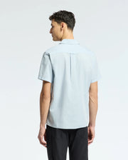 A person with short dark hair is shown from behind, wearing a light blue Psycho Bunny MENS COBAR WOVEN SHIRT - B6Q522F200 and black pants. The cooling linen blend fabric of the shirt contrasts with the plain white background.