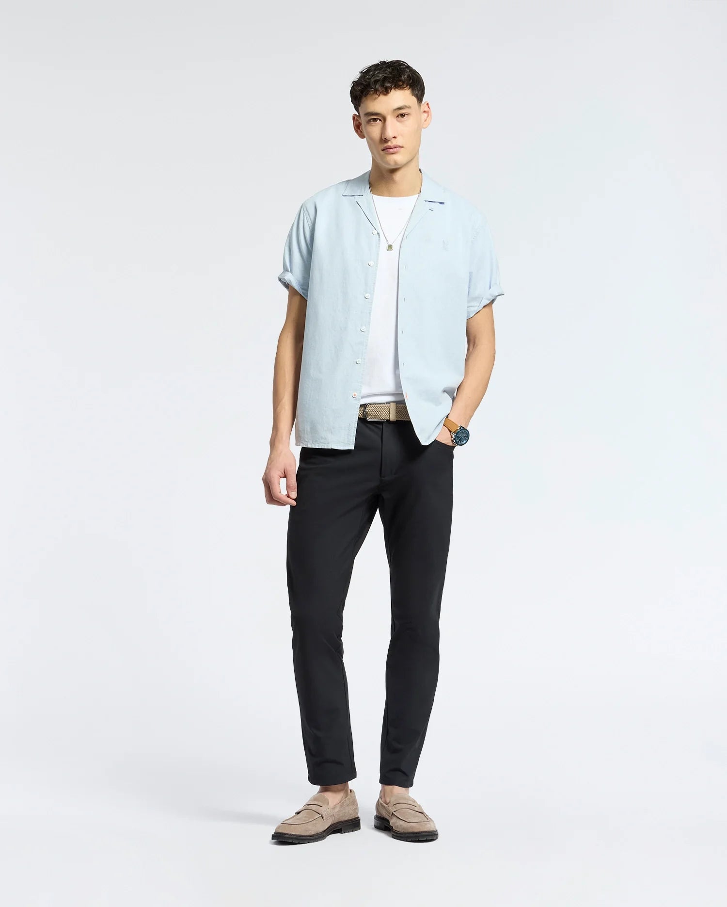 Against a plain background, a person effortlessly captures the beachwear vibe in Psycho Bunny's MENS COBAR WOVEN SHIRT - B6Q522F200, a linen blend light blue short-sleeve button-up over a white T-shirt. Paired with black pants, brown loafers, their short curly hair and watch complete the look.