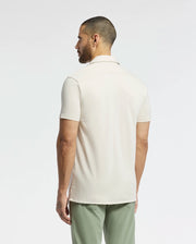 A man with a short beard, facing away, is wearing the light beige Psycho Bunny Men's Bryan Camp Collar Short Sleeve Shirt (B6Q581F200) and green pants against a plain white background.