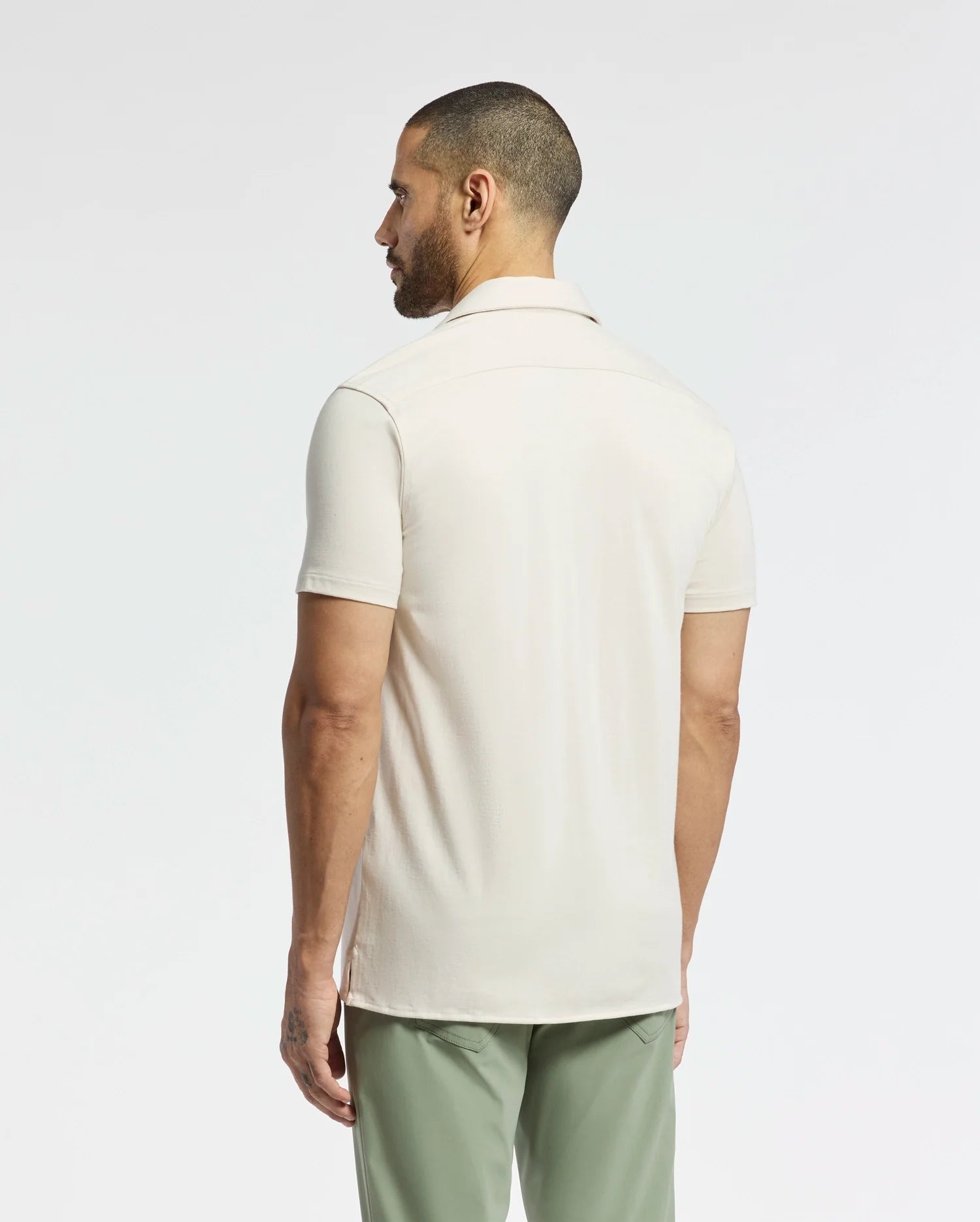 A man with a short beard, facing away, is wearing the light beige Psycho Bunny Men's Bryan Camp Collar Short Sleeve Shirt (B6Q581F200) and green pants against a plain white background.