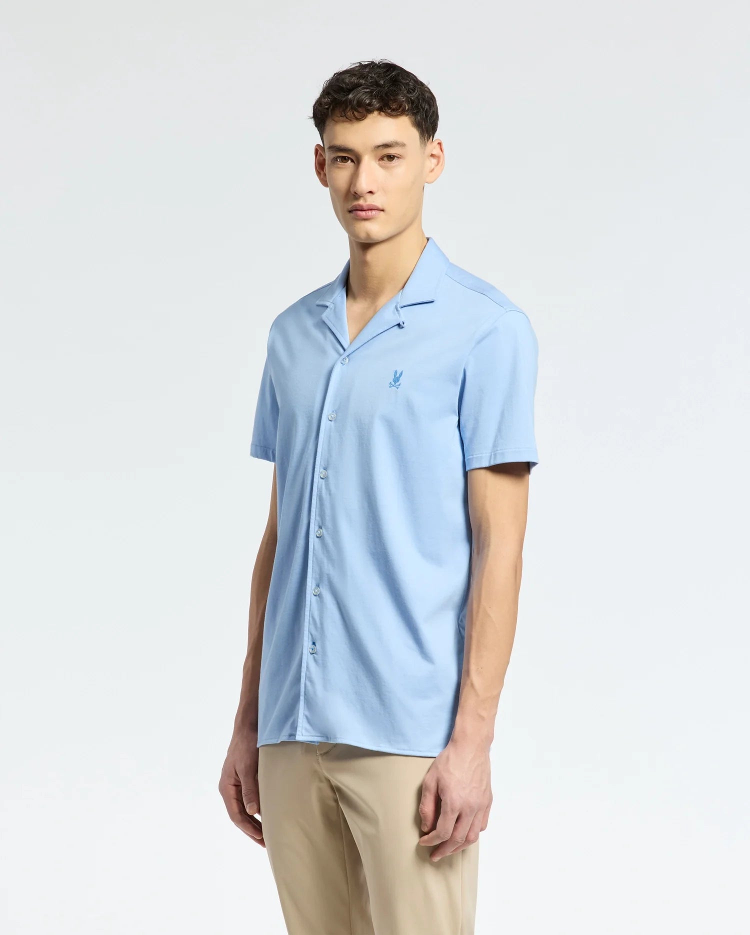 A person stands against a plain background wearing the Psycho Bunny MENS BRYAN CAMP COLLAR SHORT SLEEVE SHIRT - B6Q581F200 in light blue, featuring performance fabric and an embroidered chest design, paired with beige pants.