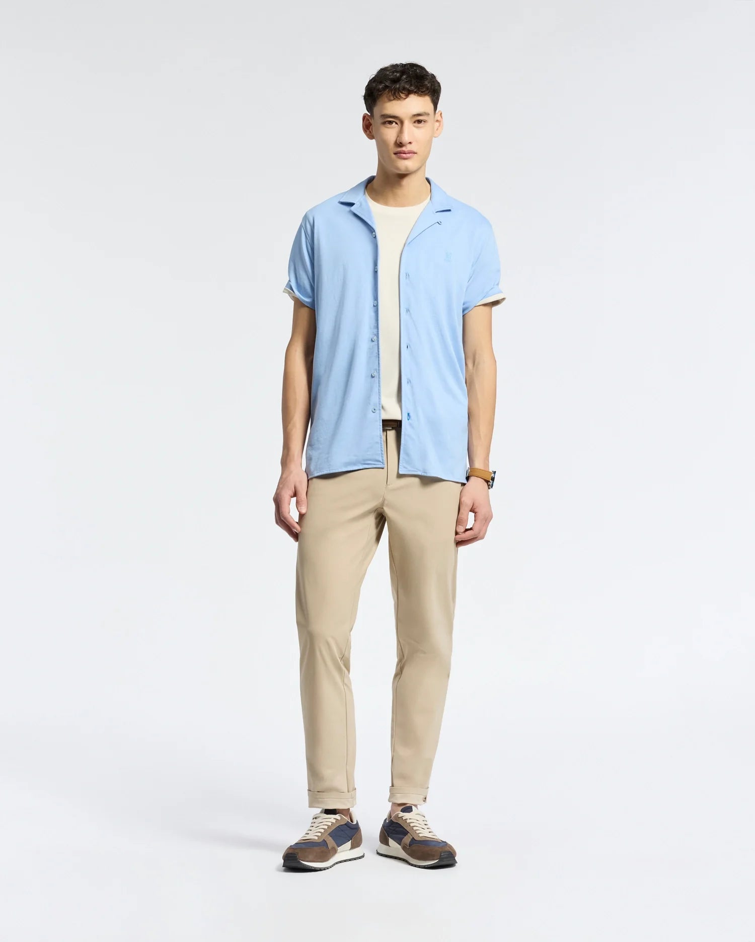 A man stands against a plain backdrop, wearing Psycho Bunny's MENS BRYAN CAMP COLLAR SHORT SLEEVE SHIRT in light blue over a white T-shirt, beige pants, and sneakers. His hands rest by his sides as he gazes slightly left.