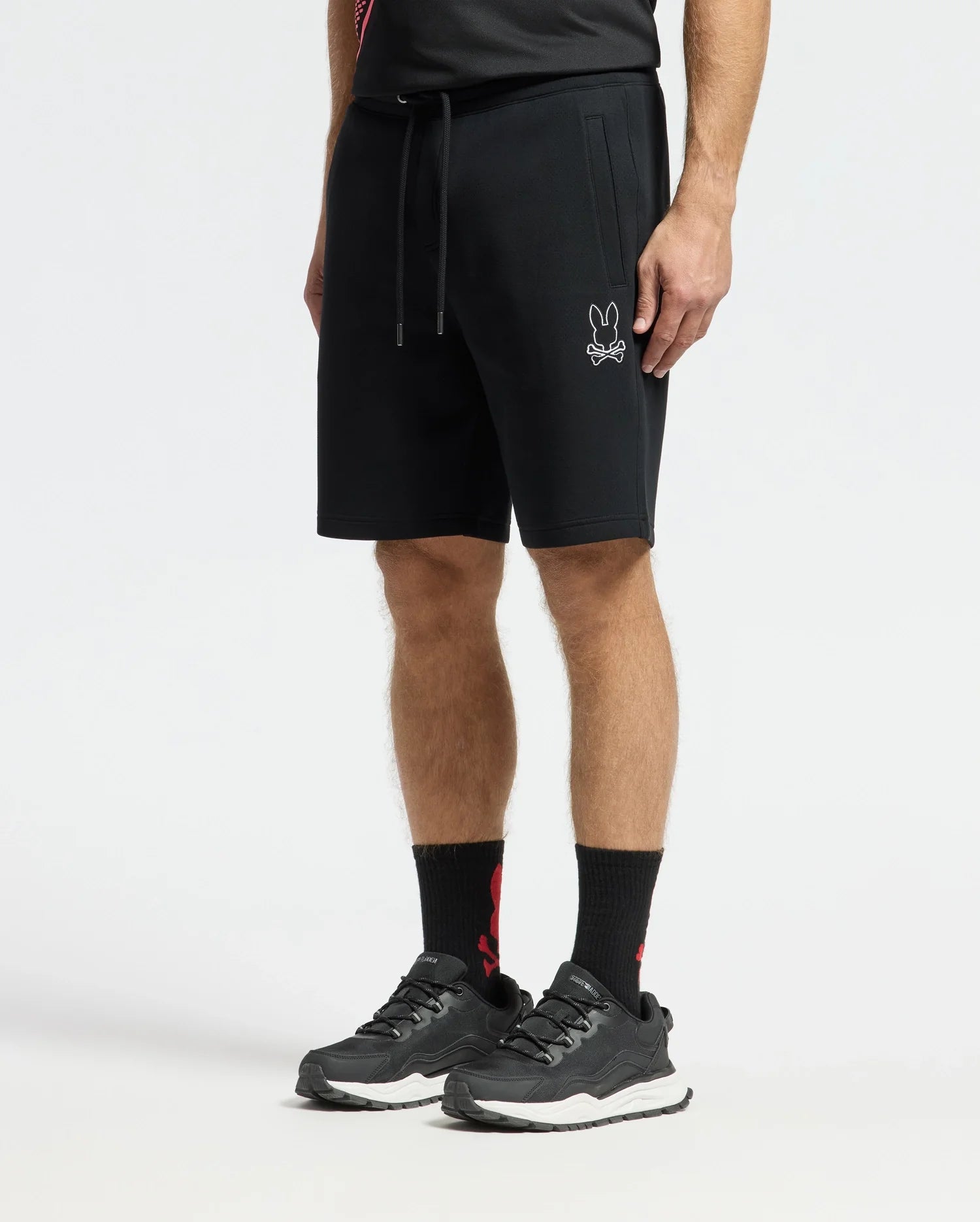 A man is wearing Psycho Bunny's MENS DACANO PERFORATED SWEATSHORT - B6R286E200 with a rabbit logo, styled for an athletic fit. He's also in black sneakers, high black socks, and a performance-oriented cotton-blend jersey shirt, all set against a plain white background.