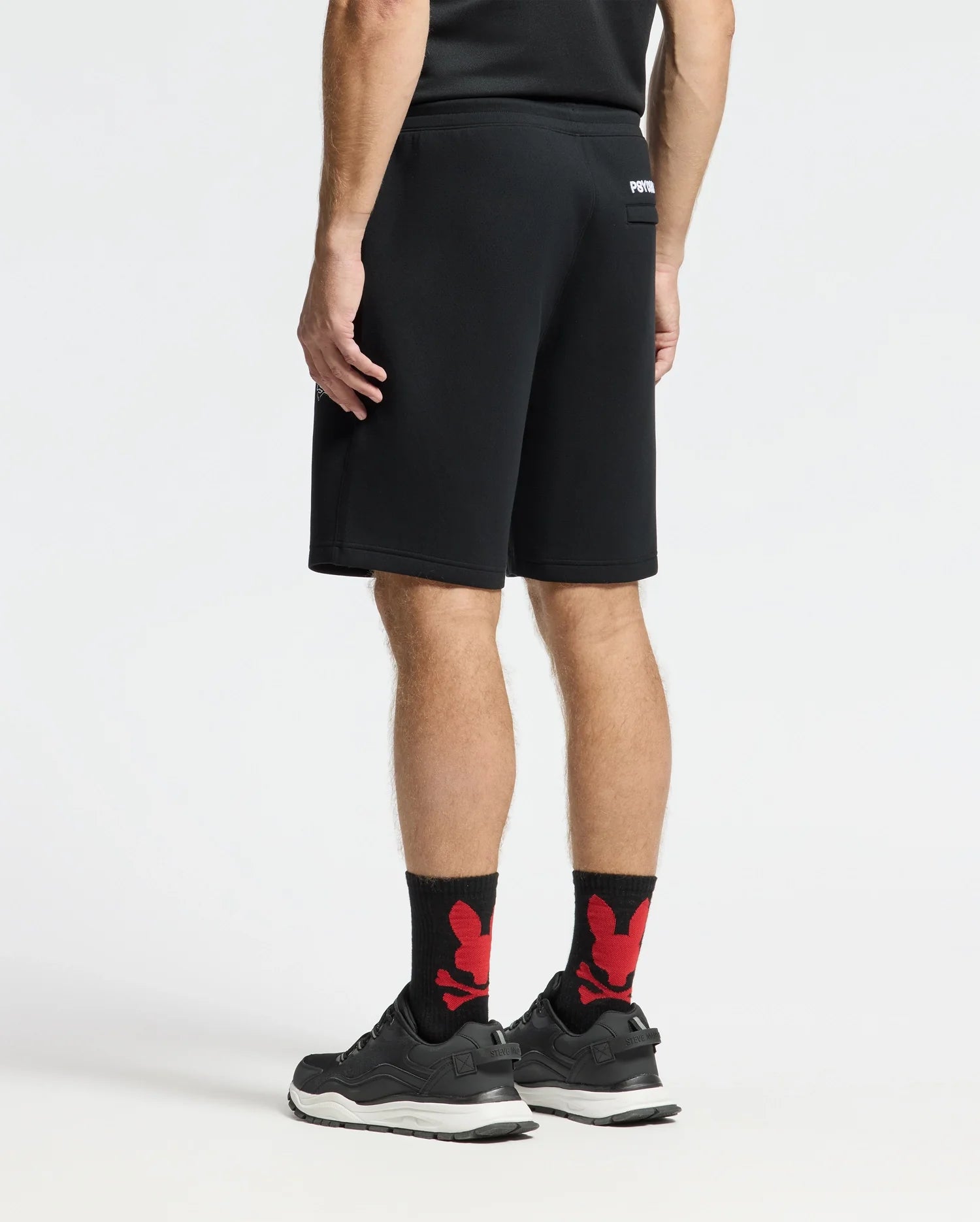 A person stands facing away, wearing a black athletic shirt and MENS DACANO PERFORATED SWEATSHORT by Psycho Bunny along with black sneakers and black socks featuring red designs. This cotton-blend jersey ensemble is performance-oriented against a plain white background.