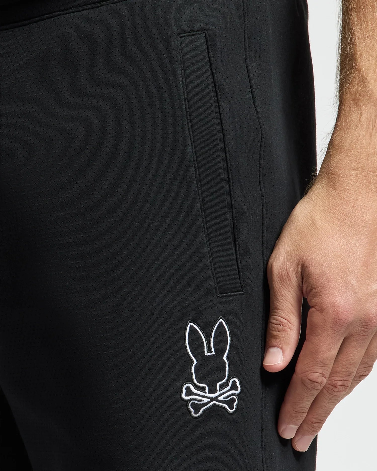 Close-up of someone wearing black MENS DACANO PERFORATED SWEATSHORT by Psycho Bunny. The cotton-blend jersey features a white bunny head with crossed bones logo near the knee. Their hand rests by their side, exuding casual confidence.