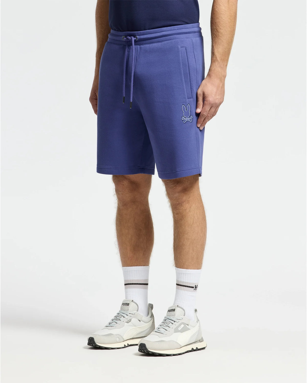 Wearing Psycho Bunny's Dacano Perforated Sweatshort in a cotton blend with side logo, someone pairs these blue drawstring shorts with a navy shirt, white socks, and sleek white sneakers featuring gray accents against a plain white backdrop.