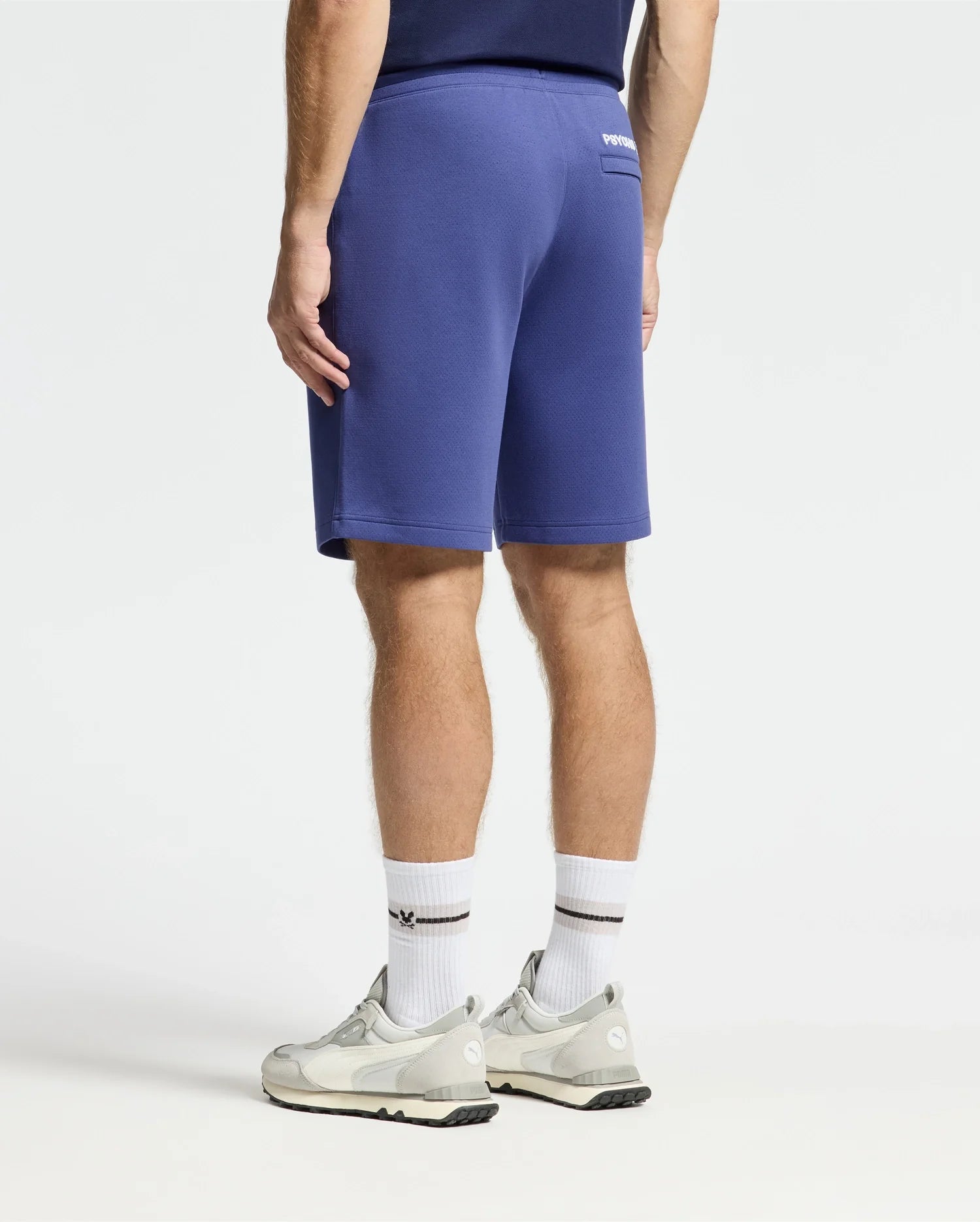 A person faces away from the camera, wearing Psycho Bunny's Dacono Perforated Sweatshorts and a navy performance cotton-blend shirt, paired with white socks and gray sneakers, creating an athletic look against a plain white background.
