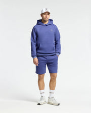 A person poses against a plain backdrop in Psycho Bunny's blue performance-oriented cotton blend hoodie and MENS DACANO PERFORATED SWEATSHORTS, styled with white socks, sneakers, and cap. The athletic fit exudes a casual, sporty vibe.