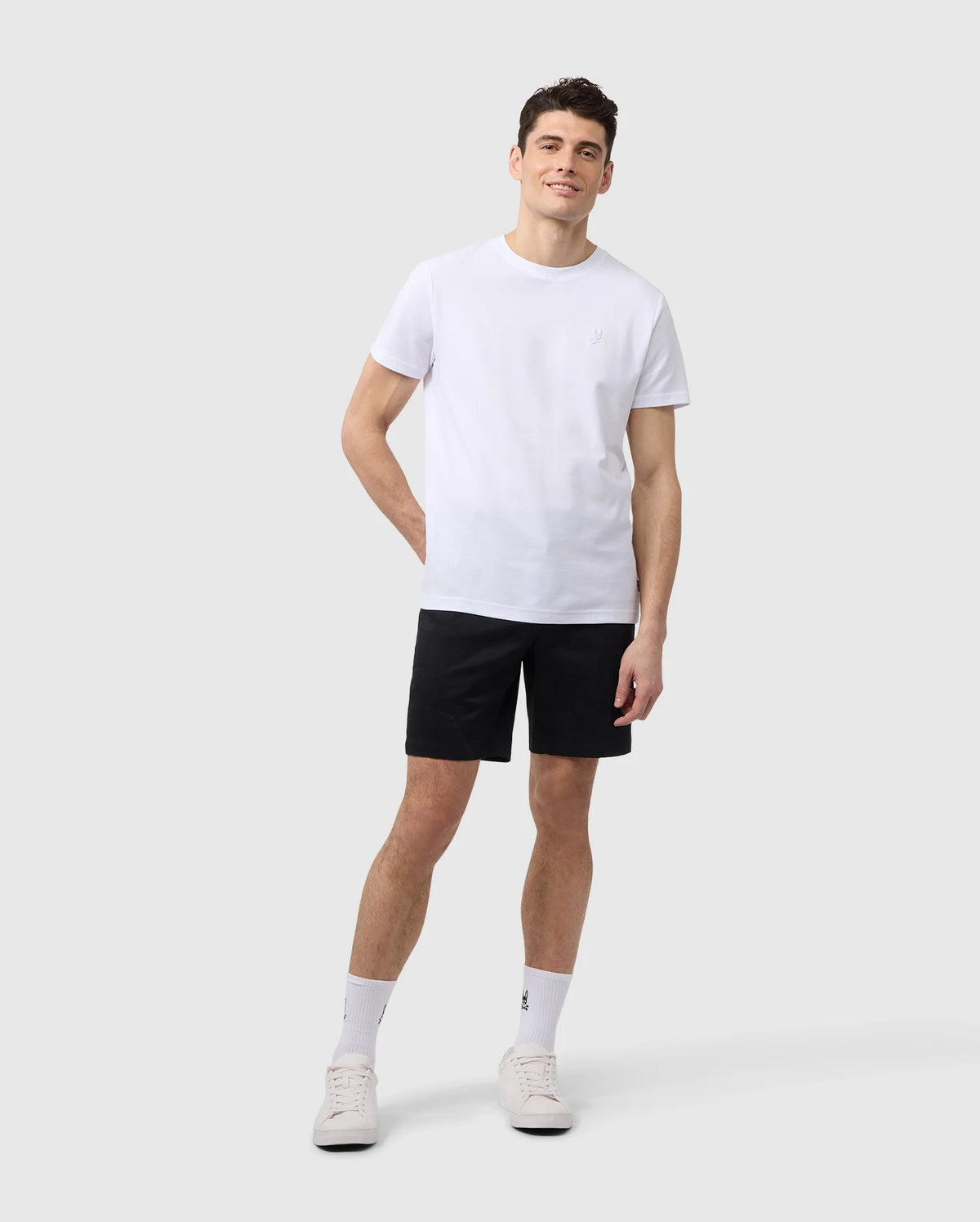 A young man wearing a plain white t-shirt, black MENS YORK CHINO SHORT - B6R357B200 shorts, white socks, and white sneakers stands against a plain light background, looking slightly to his left with a soft smile.