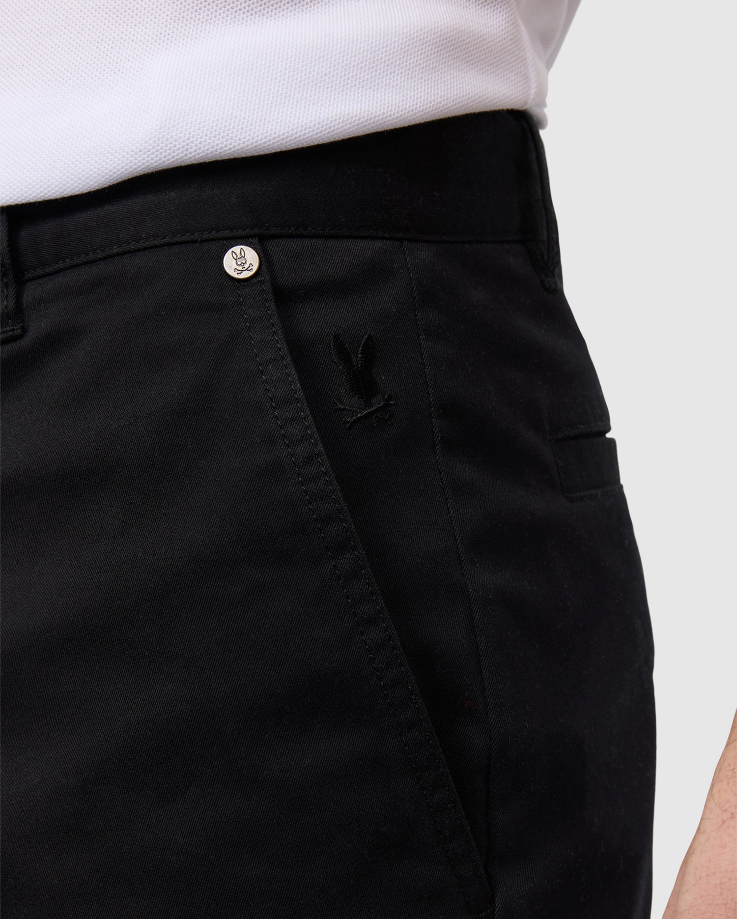 Close-up view of a person wearing black Psycho Bunny men's York chino shorts, featuring a small embroidered logo near the pocket with a visible branded button on the waistband.