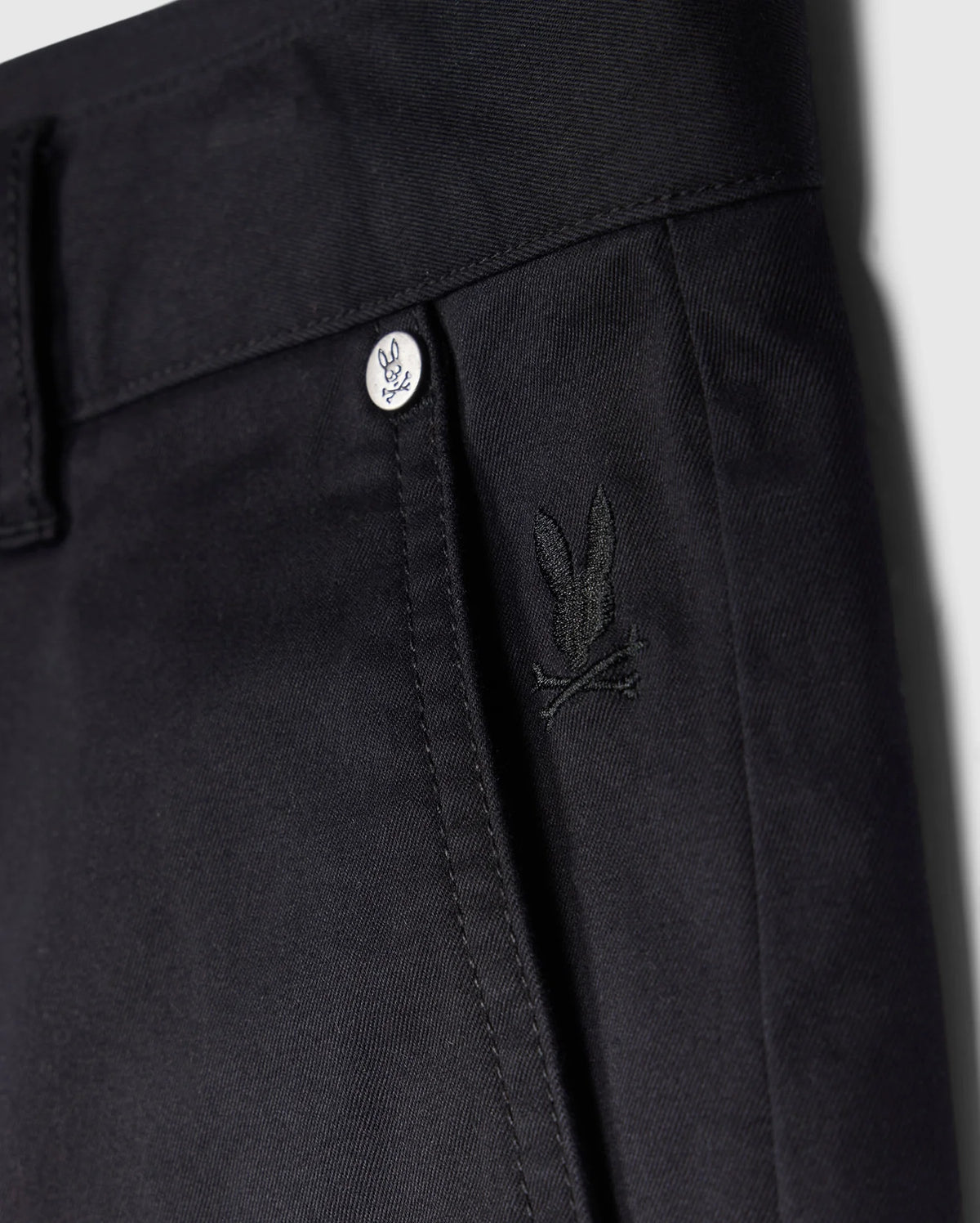 Close-up view of a black trouser waistline featuring a button with the Psycho Bunny logo and a small embroidered Psycho Bunny logo on the front pocket of men's MENS YORK CHINO SHORT - B6R357B200.