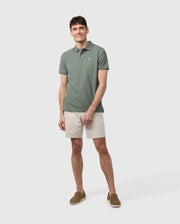 A young man smiling, standing against a white background, wearing a light green polo shirt, beige Psycho Bunny Men's York Chino Shorts, and brown loafers.