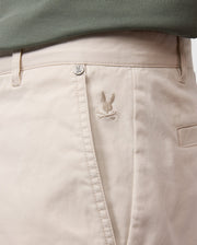 Close-up of light beige Psycho Bunny men's York chino shorts with a small, embroidered bird logo near the right front pocket, partially covered by a teal fabric edge.
