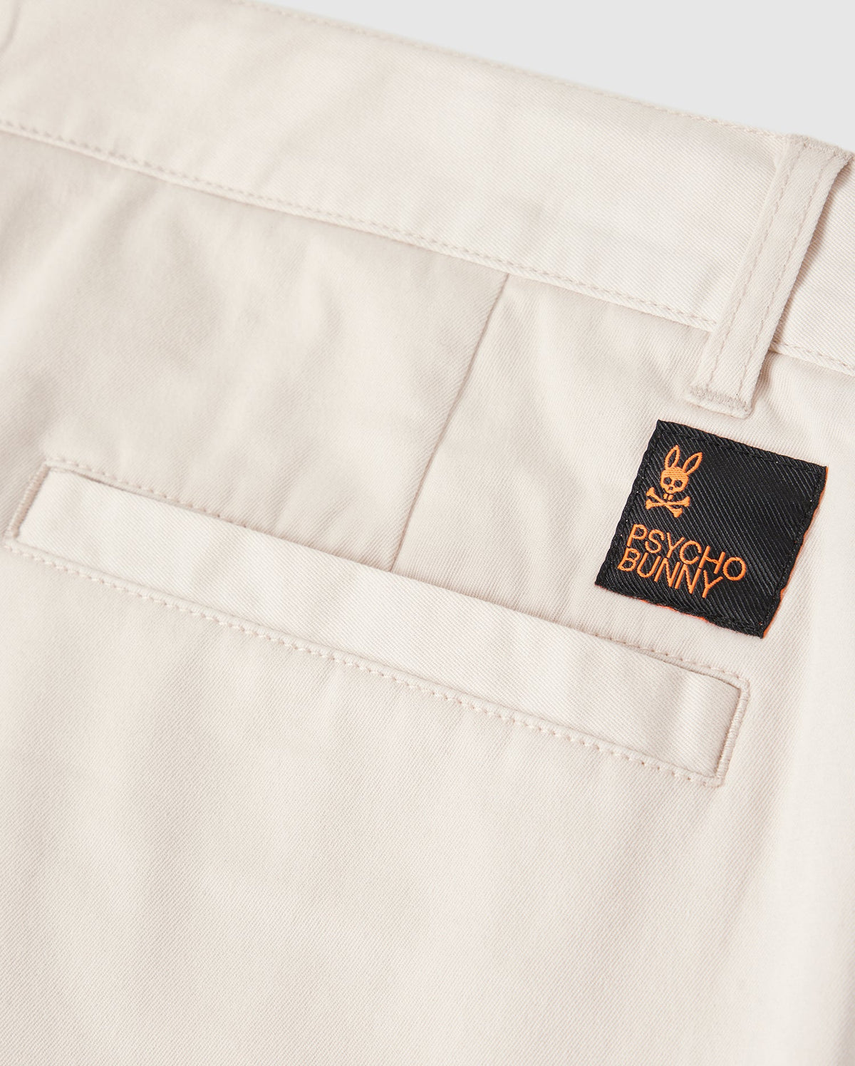 Close-up view of Psycho Bunny men's chino shorts in beige with a black Psycho Bunny brand logo patch on them. The patch features a stylized rabbit skull and crossbones design.