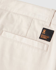 Close-up view of Psycho Bunny men's chino shorts in beige with a black Psycho Bunny brand logo patch on them. The patch features a stylized rabbit skull and crossbones design.
