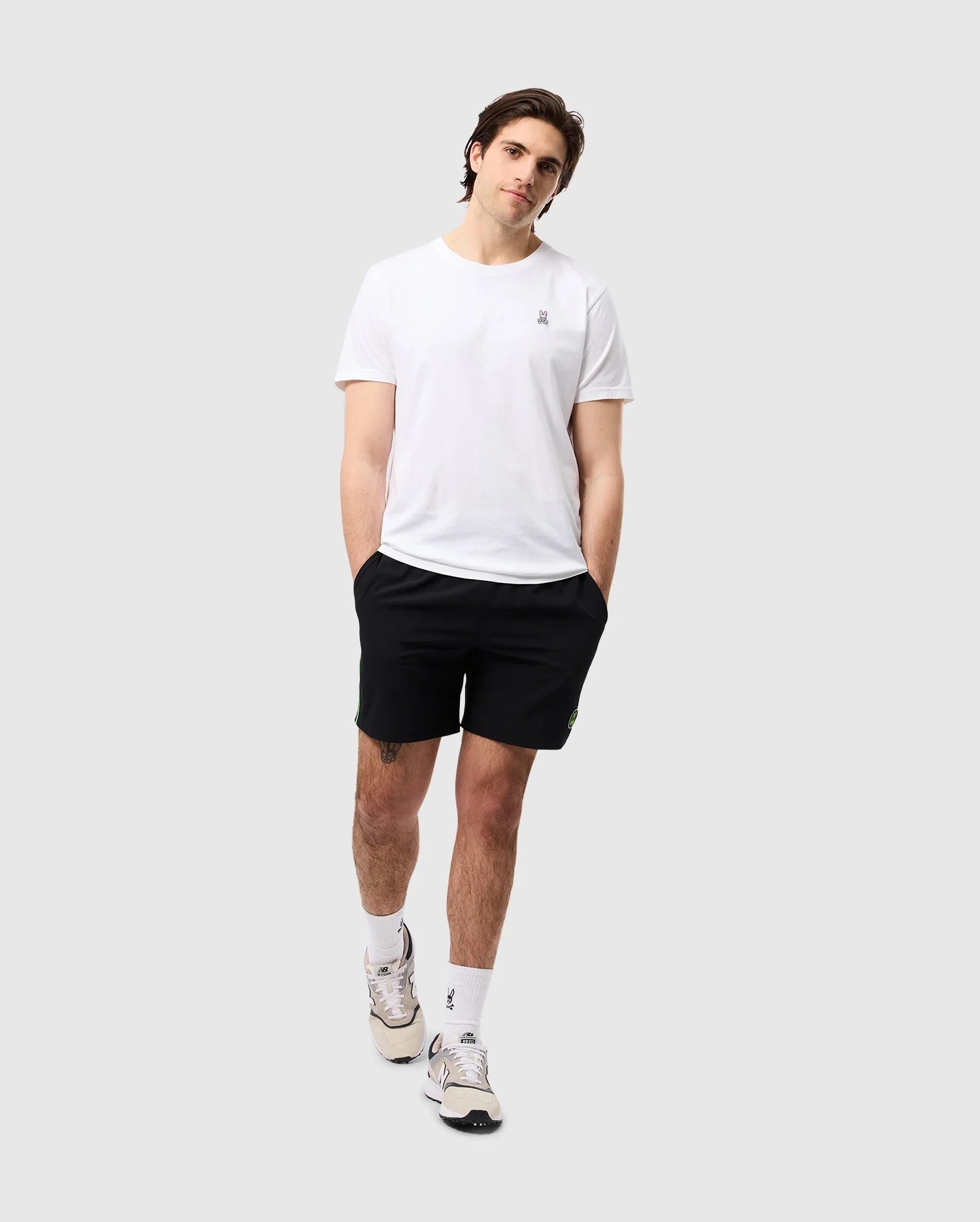 A person wearing a plain white t-shirt, Psycho Bunny MENS SONTERRA STRIPE SPORT SHORT - B6R464C200 made of quick-drying, breathable fabric, and white sneakers with socks stands casually with hands in pockets against a light-gray background. The person's head is slightly tilted to one side, and they have a relaxed expression.