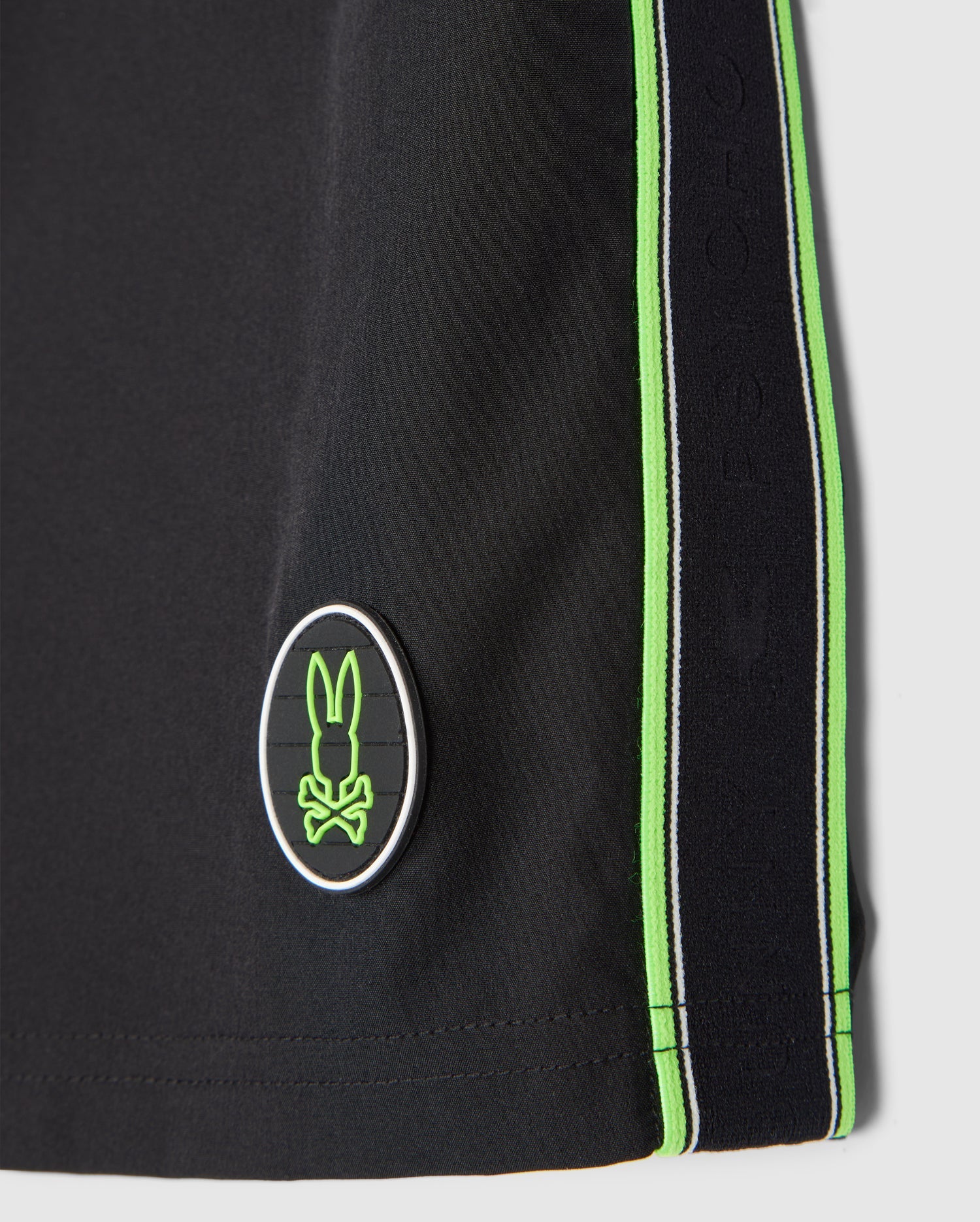 Close-up of a black Psycho Bunny MENS SONTERRA STRIPE SPORT SHORT - B6R464C200 featuring vertical green stripes along the side. There's an oval emblem with a stylized rabbit logo in green and white near the bottom hem. The quick-drying, breathable fabric showcases detailed stitching, enhancing both comfort and durability.