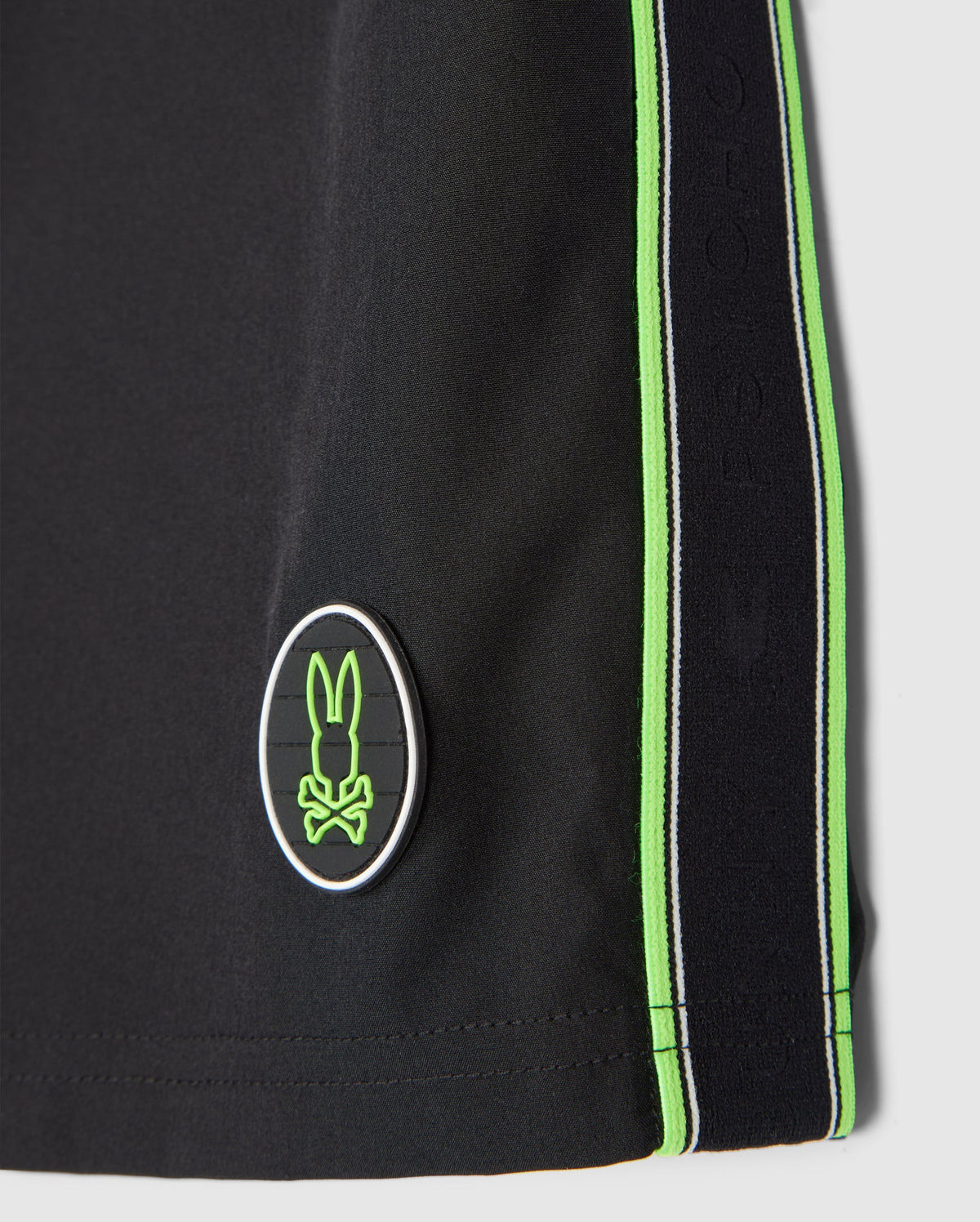 Close-up of a black Psycho Bunny MENS SONTERRA STRIPE SPORT SHORT - B6R464C200 featuring vertical green stripes along the side. There's an oval emblem with a stylized rabbit logo in green and white near the bottom hem. The quick-drying, breathable fabric showcases detailed stitching, enhancing both comfort and durability.