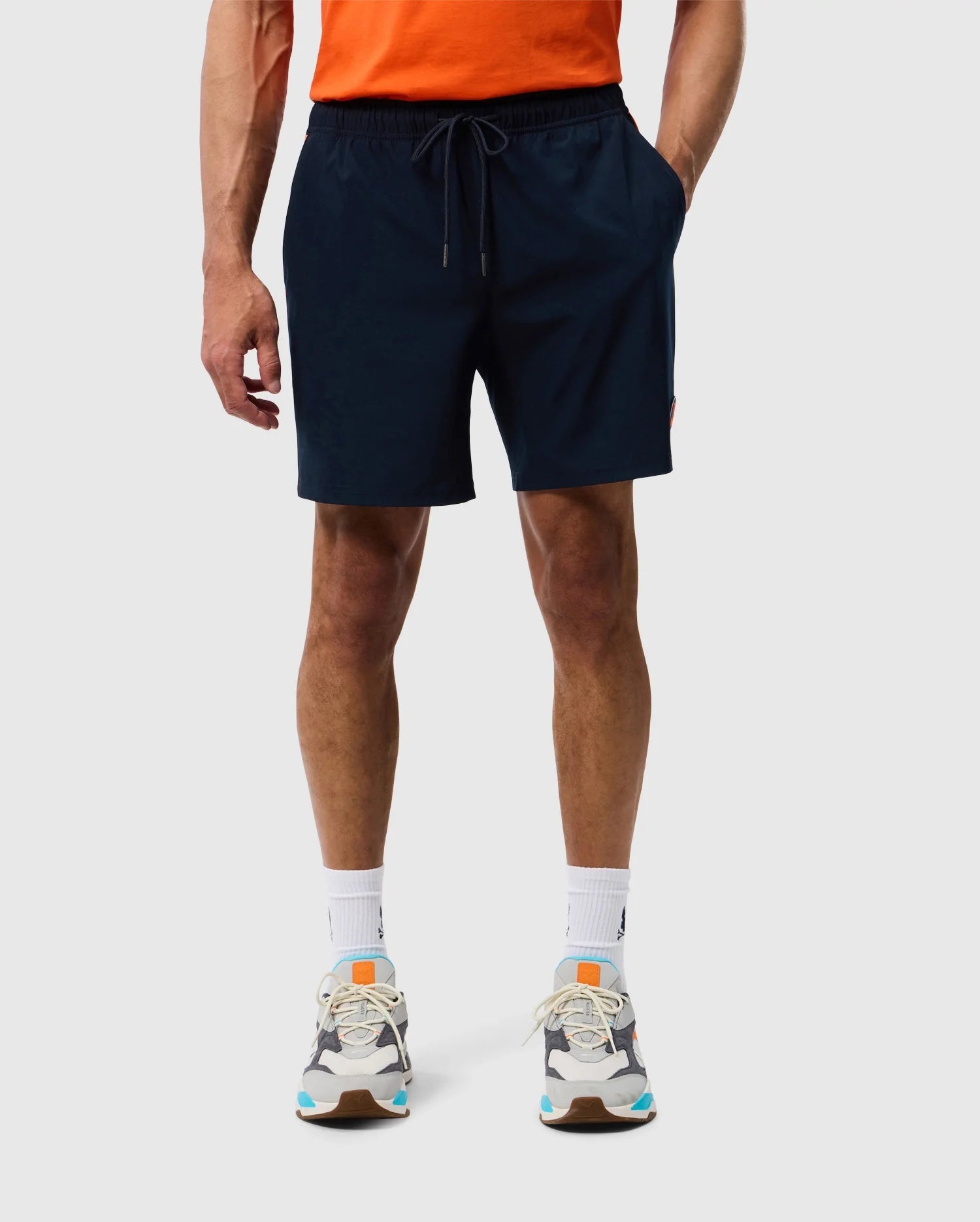 A person is standing against a plain background, wearing a bright orange t-shirt, dark blue Psycho Bunny MENS SONTERRA STRIPE SPORT SHORT - B6R464C200 with a drawstring made from stretch fabric, white socks, and multicolored athletic shoes. The individual is visible from the shoulders down.