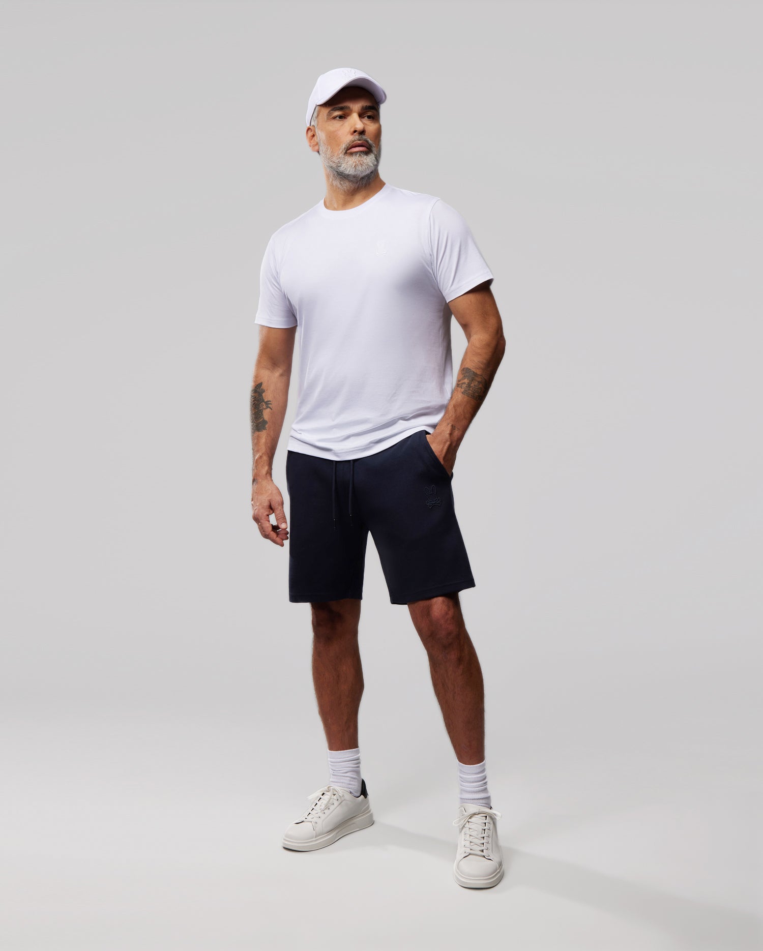 A man stands against a plain background wearing a white cap, white t-shirt, and navy blue MENS OUTLINE SWEATSHORTS - B6R507ARCN from Psycho Bunny. He has white socks and sneakers, one hand in his pocket, the other arm by his side. Sporting a beard and mustache, he gazes slightly off-camera.