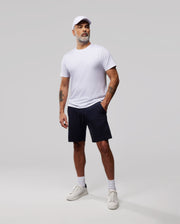 A man stands against a plain background wearing a white cap, white t-shirt, and navy blue MENS OUTLINE SWEATSHORTS - B6R507ARCN from Psycho Bunny. He has white socks and sneakers, one hand in his pocket, the other arm by his side. Sporting a beard and mustache, he gazes slightly off-camera.