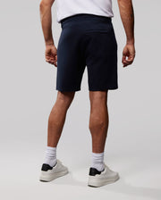 A person stands facing away, wearing a white Psycho Bunny T-shirt, dark navy Psycho Bunny MENS OUTLINE SWEATSHORTS - B6R507ARCN, white socks, and white sneakers with black accents. The background is plain and light gray.