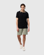 A young man in a black polo shirt made of honeycomb fabric and light green MENS SHIRO KNIT HONEYCOMB SHORT - B6R567C200 shorts stands against a white background, wearing white sneakers. He looks directly at the camera with a neutral expression.