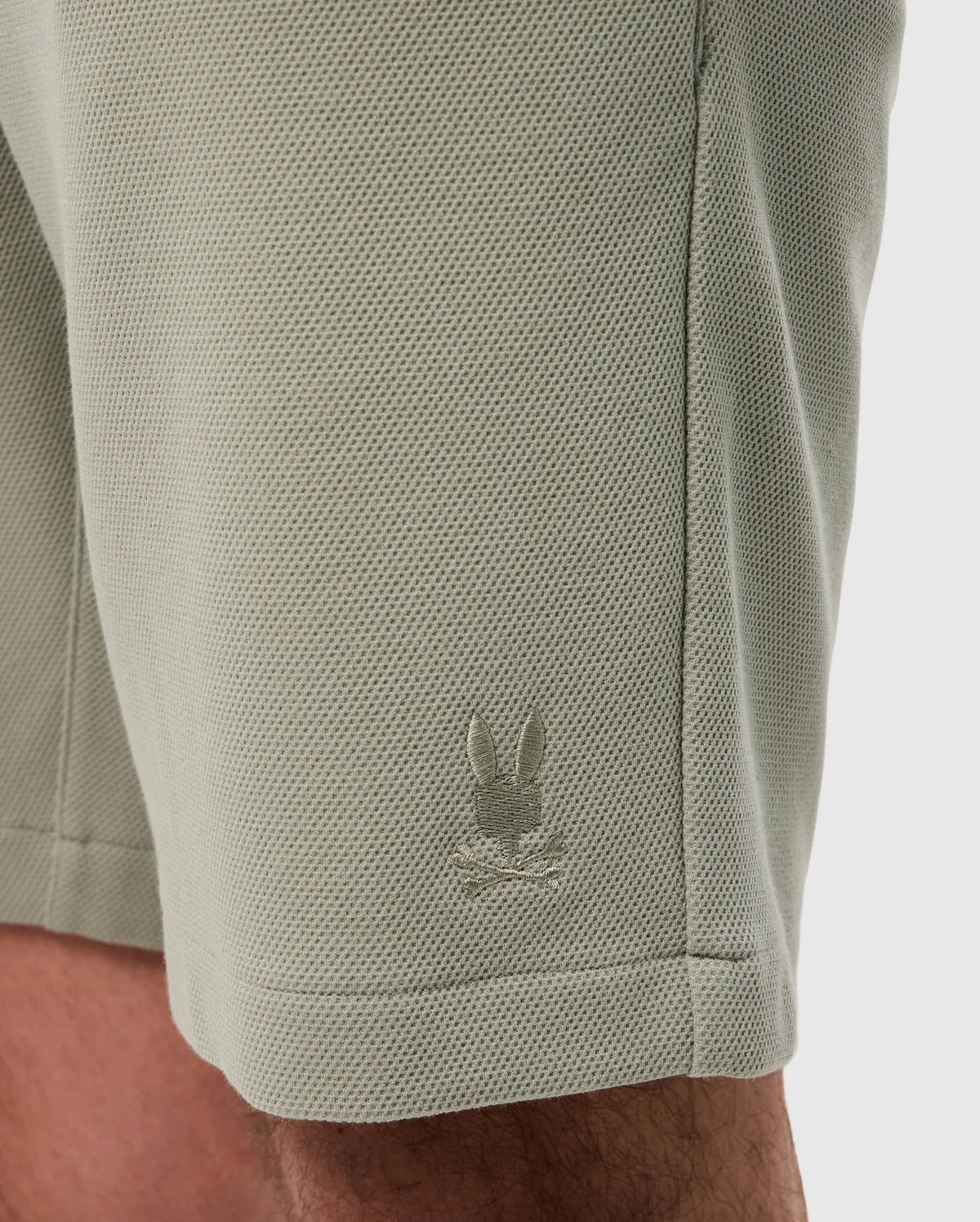 Close-up of a light green, sweat-wicking fabric with a small, white embroidered logo of a rabbit on the lower side near a seam on the Psycho Bunny MENS SHIRO KNIT HONEYCOMB SHORT - B6R567C200.