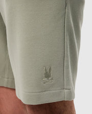 Close-up of a light green, sweat-wicking fabric with a small, white embroidered logo of a rabbit on the lower side near a seam on the Psycho Bunny MENS SHIRO KNIT HONEYCOMB SHORT - B6R567C200.