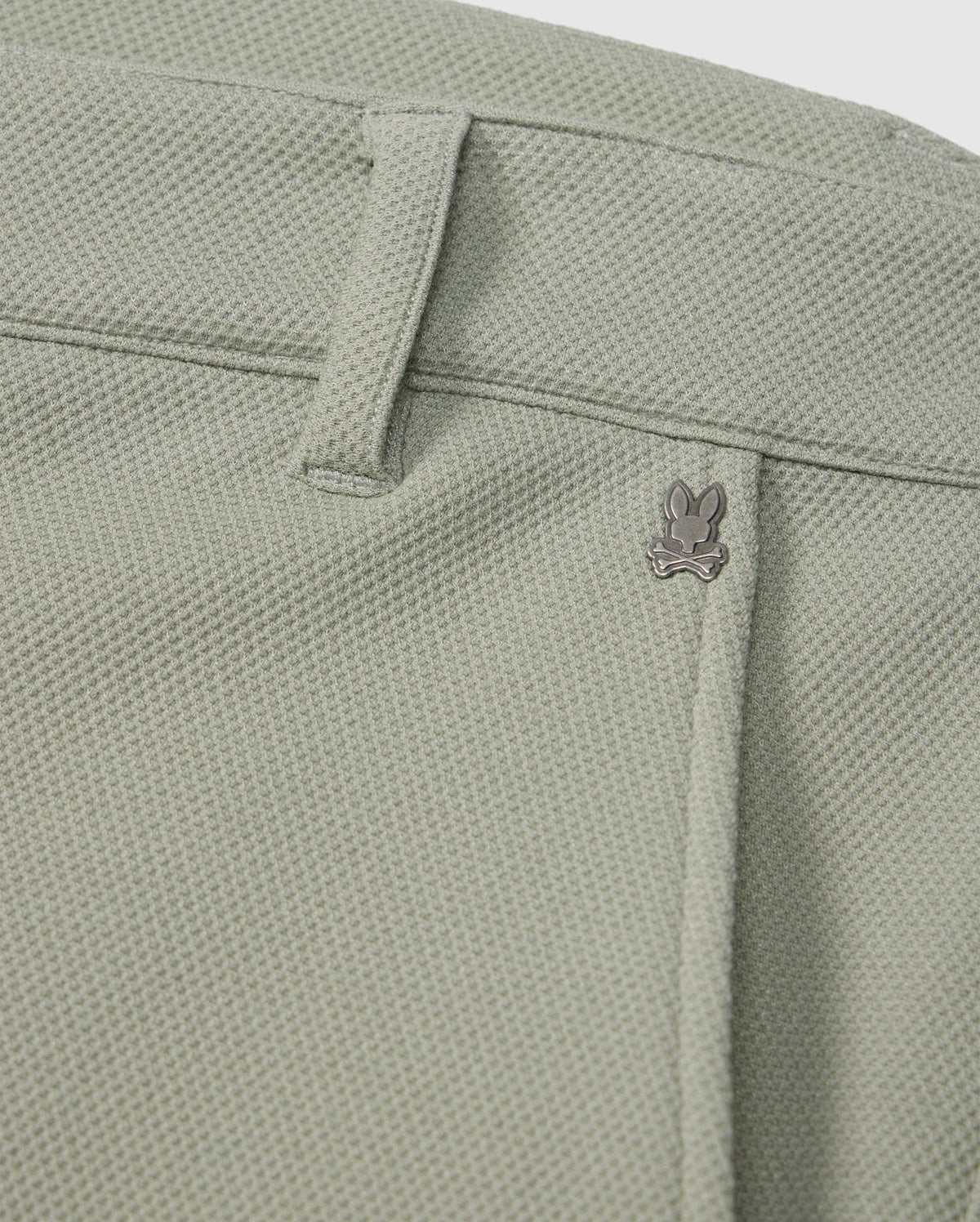 Close-up of a light green Psycho Bunny honeycomb fabric with a subtle texture, featuring a small metal bunny logo near the seam of a pocket.
