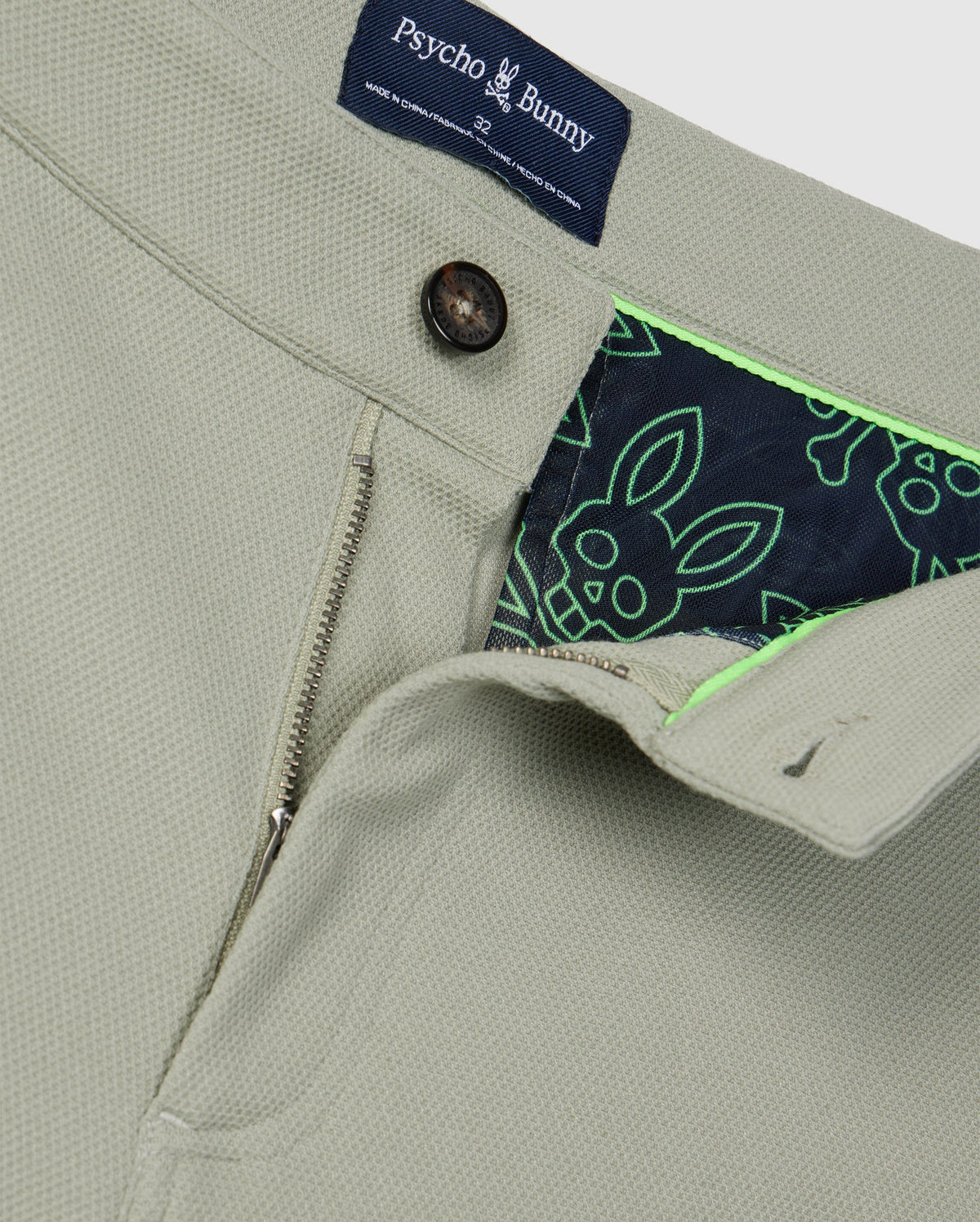 Close-up of a beige Psycho Bunny MENS SHIRO KNIT HONEYCOMB SHORT - B6R567C200 jacket with a partially unzipped front, revealing a vibrant honeycomb fabric lining patterned with green and navy blue bunnies. The collar tag and front button are also visible.