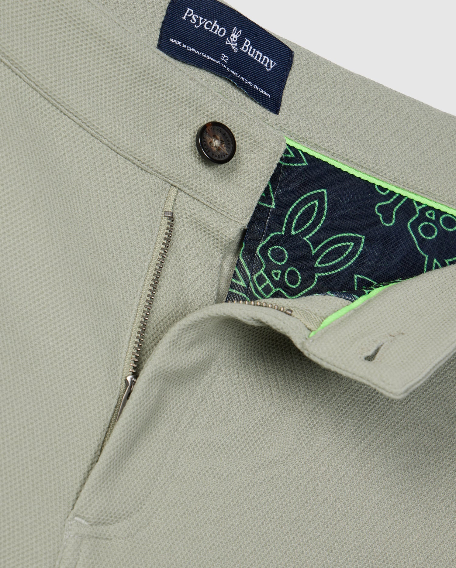Close-up of a beige Psycho Bunny MENS SHIRO KNIT HONEYCOMB SHORT - B6R567C200 jacket with a partially unzipped front, revealing a vibrant honeycomb fabric lining patterned with green and navy blue bunnies. The collar tag and front button are also visible.