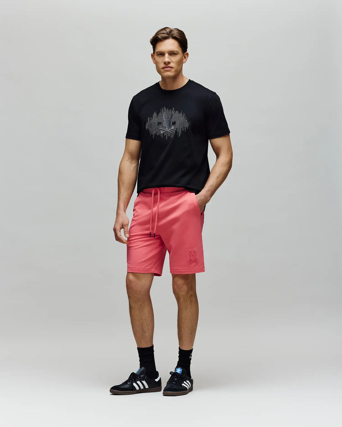 A man with short brown hair and a neutral expression wears a black graphic tee, Psycho Bunny MENS LEVANZO FRENCH TERRY SWEATSHORT - B6R578F200 in bright pink, black socks, and black Adidas sneakers against a plain background.