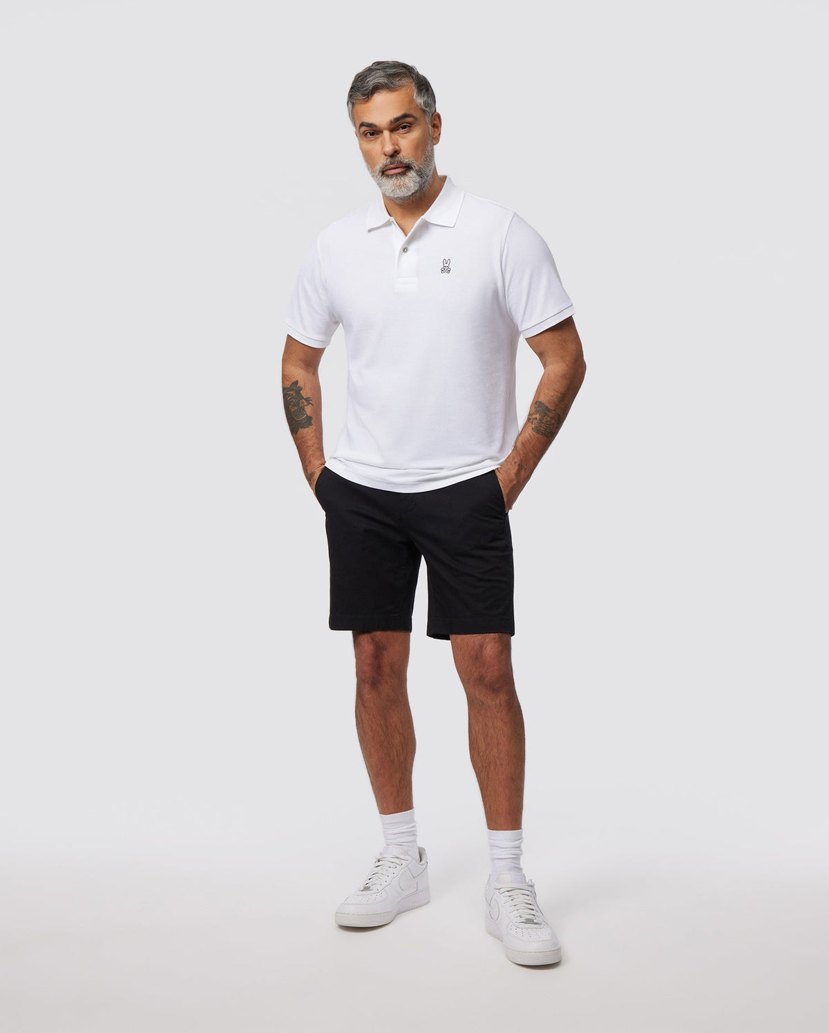 A man with short gray hair and a beard stands against a plain background, wearing a slim fit white polo shirt made of breathable cotton, black MENS HAYDEN SHORT - B6R757ARCE by Psycho Bunny, and white sneakers. His hands are in his pockets, revealing tattoos on both arms.