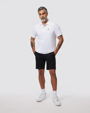 A man with short gray hair and a beard stands against a plain background, wearing a slim fit white polo shirt made of breathable cotton, black MENS HAYDEN SHORT - B6R757ARCE by Psycho Bunny, and white sneakers. His hands are in his pockets, revealing tattoos on both arms.