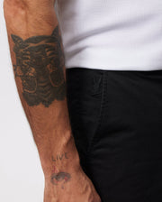 A person's arm with a large tattoo of a cat face is shown alongside Psycho Bunny MENS HAYDEN SHORT - B6R757ARCE with a small embroidered logo of a rabbit near the pocket. The person is wearing a white shirt made of breathable cotton. Their wrist has another tattoo that reads "LIVE" above a small eye design.