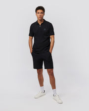 A man stands against a plain white background wearing a black polo shirt and Psycho Bunny MENS GABLE REGULAR FIT SPORT SHORT - B6R761ARCN. He has short, curly hair and is looking directly at the camera with his hands in his hidden zip pocket. He is also wearing white socks and white sneakers.