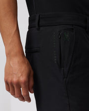 Close-up of a person wearing black pants with a small green Psycho Bunny logo and the words "PSYCHO BUNNY" vertically stitched near the pocket. The durable stretch cotton-nylon blend ensures comfort, while the person’s hand is visible resting alongside the hidden zip pocket. The background is plain and white. They are wearing MENS GABLE REGULAR FIT SPORT SHORT - B6R761ARCN by Psycho Bunny.