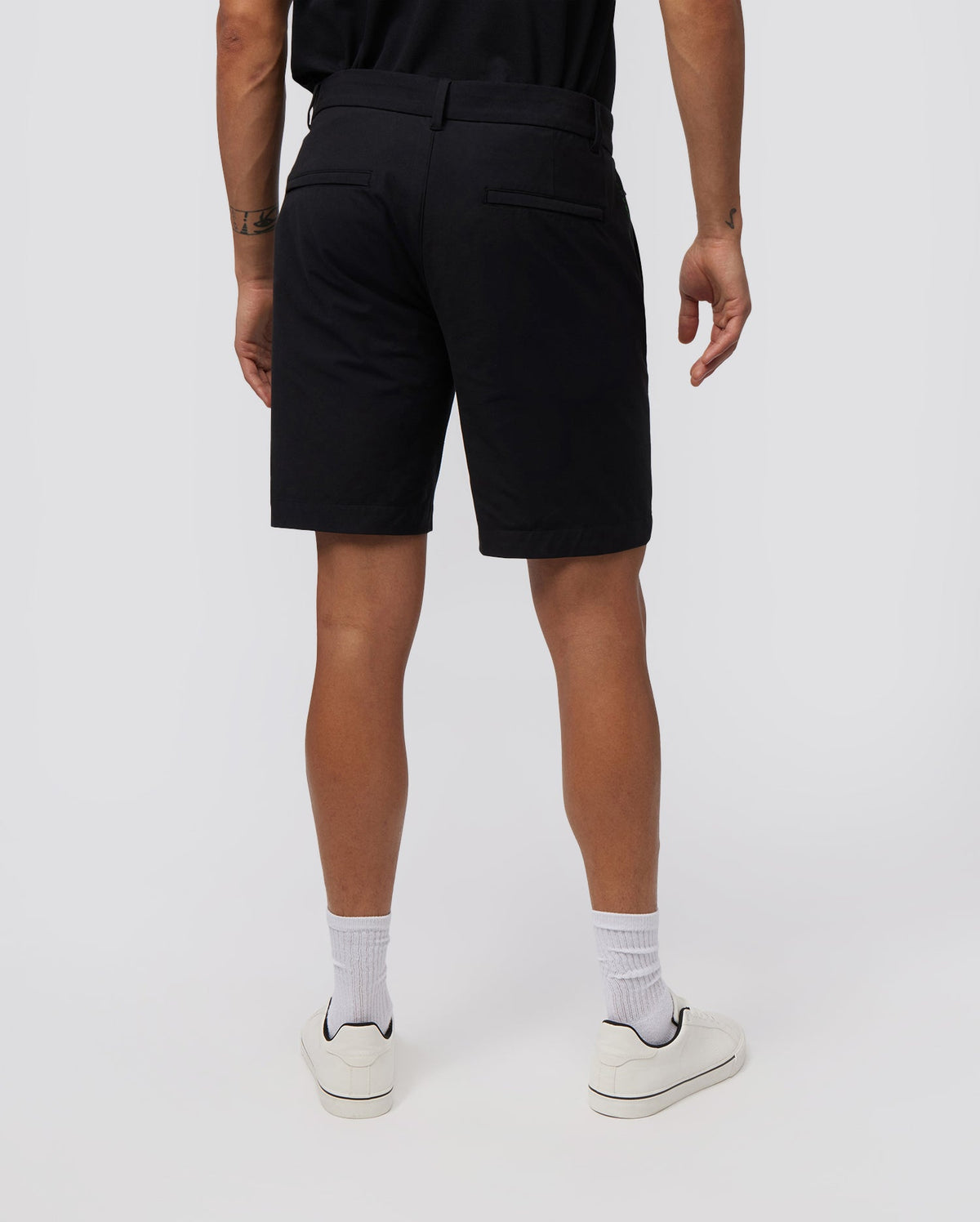 A person wearing black Psycho Bunny MENS GABLE REGULAR FIT SPORT SHORT - B6R761ARCN and a black shirt is standing with their back to the camera. They are also sporting white socks and white sneakers. The background is plain and light-colored. These durable stretch cotton-nylon shorts feature a hidden zip pocket for convenience.