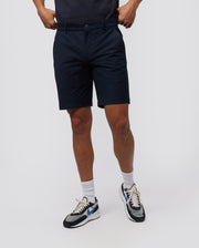A person wearing knee-length, navy blue Psycho Bunny MENS GABLE REGULAR FIT SPORT SHORT - B6R761ARCN made from durable stretch cotton-nylon, white socks, and white sneakers with blue, black, and beige accents stands against a plain white background. The individual's hands are tucked into the pockets of their shorts.